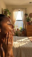 Young African American woman applying skincare product in sunlit bedroom with houseplants for morning wellness routine.