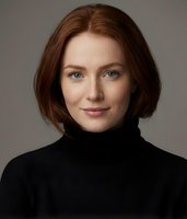 Professional portrait of confident young Caucasian woman with auburn hair wearing black turtleneck sweater against neutral background for business use.