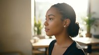Young African American businesswoman in profile looking thoughtfully out office window during golden hour lighting in modern workplace setting.