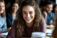 Young Caucasian woman with curly hair and genuine smile in casual burgundy sweater sitting at cafe table, expressing joy and positive emotions during social gathering.