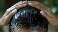 Close-up of hands with manicured nails applying hair product on wet, slicked back dark hair against blurred green background.