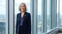 Professional mature Caucasian businesswoman with gray bob haircut in navy suit standing confidently in modern office with city skyline view through windows.
