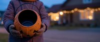 Person holding carved pumpkin jack-o-lantern at twilight with warm house lights in background during Halloween season celebration.