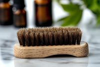 Wooden beard brush with natural horsehair bristles on marble surface, essential oil bottles and green leaves in background, grooming accessory for men.