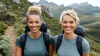 Two diverse female hikers with bright smiles wearing backpacks and athletic wear on mountain trail, enjoying outdoor adventure against scenic alpine landscape.