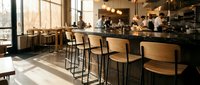 Modern cafe interior with wooden bar stools at counter, natural light from large windows, staff working behind espresso bar, contemporary restaurant design for hospitality businesses.