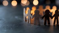 Paper cutout figures holding hands in unity against blurred warm bokeh lights background, symbolizing community, teamwork and social connection.