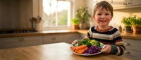 Happy young Caucasian boy smiling at kitchen table with colorful plate of fresh vegetables including broccoli, carrots and purple cabbage for healthy eating.