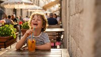 Happy young boy with curly blonde hair enjoying orange juice at outdoor cafe terrace, smiling and laughing during summer vacation in Mediterranean setting.