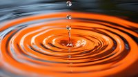 Water drops falling into orange liquid creating concentric ripple patterns with splash and reflection against dark background. Macro photography captures fluid dynamics.