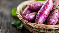 Fresh purple sweet potatoes in woven basket on rustic wooden surface with green leaves, macro food photography showing natural organic produce.