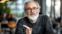 Senior Caucasian man with gray beard and glasses wearing dark sweater smiling warmly while gesturing during conversation in modern cafe setting with blurred background.