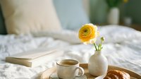 Single yellow ranunculus flower in ceramic vase with morning coffee cup and croissant on white bedding creating cozy breakfast in bed atmosphere.