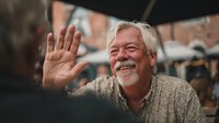 Cheerful senior Caucasian man with white beard waving hello outdoors, smiling warmly at camera in casual sweater for greeting and communication concepts.