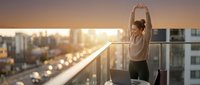 Woman stretching arms overhead on high-rise balcony at sunrise, urban skyline background, wellness and morning routine concept for lifestyle and health content.