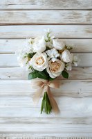 Elegant bridal bouquet with cream roses and white flowers tied with beige ribbon, displayed on rustic whitewashed wooden background.