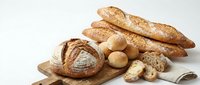 Assorted fresh baked bread loaves and rolls arranged on wooden board with linen cloth, including baguettes, sourdough, and round artisan loaves.