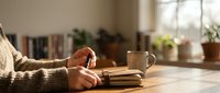 Cozy reading moment with hands holding open book on wooden table in warm sunlit room with plants and bookshelves creating peaceful atmosphere for relaxation.