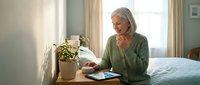 Senior Caucasian woman with gray hair using digital tablet while sitting on bed in bright bedroom, morning light streaming through window.