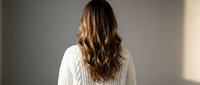 Young woman with long wavy brown hair wearing white knit sweater standing with back turned in soft natural lighting for lifestyle and fashion content.