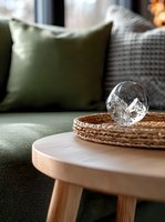 Overturned crystal glass on woven rattan coaster atop wooden side table, with green cushions and textured throw in cozy living room setting. Soft natural lighting.