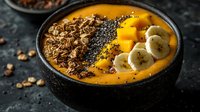 Healthy mango smoothie bowl topped with granola, chia seeds, banana slices and nuts for nutritious breakfast meal on dark slate background.