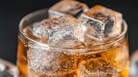 Cold carbonated drink with ice cubes in glass on dark background, refreshing cola beverage with bubbles and fizz in macro close up view.