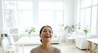 Young woman with dark hair and medium skin tone smiling in bright, minimalist home office with large windows, potted plants, and modern furniture for lifestyle, wellness, and workspace concepts.