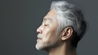 Senior Asian man with gray hair and beard in profile view against dark background showing peaceful serene expression with eyes closed, wellness concept.