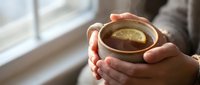 Hands holding warm ceramic mug with lemon tea by window, cozy comfort beverage for relaxation and wellness.