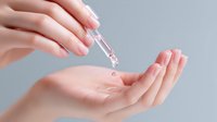 Female hands applying transparent serum or hyaluronic acid from dropper, skincare routine against gray background.