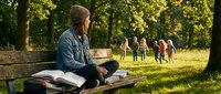Young woman reading book on park bench while friends play in background during golden hour sunset with warm natural lighting and green trees.
