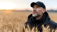 Middle-aged Caucasian farmer in black cap and jacket standing in golden wheat field at sunset, agricultural worker contemplating harvest season.