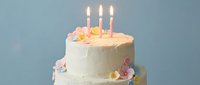 White frosted birthday cake with colorful sugar flowers and three lit candles on blue background for celebration party dessert design.