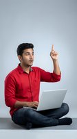 Young Indian man in red shirt pointing up with eureka moment while using laptop computer sitting cross-legged on floor against gray background.