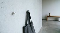 Minimalist concrete wall with modern black hook holding gray tote bag in contemporary interior space with wooden bench and polished floor.