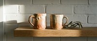Two handmade ceramic mugs with warm earthy tones rest on a wooden shelf against a white brick wall, with a sprig of green leaves nearby.
