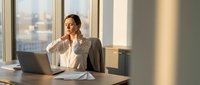 Young Caucasian businesswoman sitting at office desk with laptop, touching neck, feeling pain or stress from overwork in modern workspace.