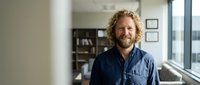 Smiling middle-aged Caucasian businessman with curly hair in casual blue shirt standing confidently in modern office workspace for professional portraits.