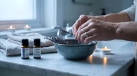 Hands preparing natural spa treatment with dried lavender in ceramic bowl surrounded by essential oil bottles and glowing candles in serene wellness setting.