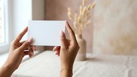 Hands holding blank white card with dried wheat and vase in soft neutral interior background for greeting, invitation, or promotional messaging.