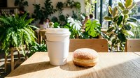 Takeaway coffee cup and pastry on wooden table with potted plants and sunlight in modern cafe setting.