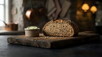 Freshly baked whole grain bread loaf with seeds and small cupcake on rustic wooden board, warm bakery setting with blurred background lighting.