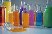 Colorful glass bottles filled with vibrant liquids in yellow, orange, purple, pink, blue and green creating rainbow laboratory setup on white surface in soft focus.