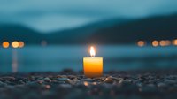 Glowing candle on pebble beach at dusk with blurred bokeh lights across water, creating peaceful meditation and wellness atmosphere for spa and relaxation designs.