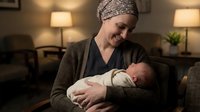 Smiling Caucasian woman wearing headscarf holds newborn baby in cozy living room with warm lighting. Motherhood, family bonding, postpartum care concept.