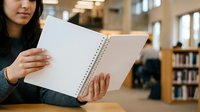 Woman with dark hair and medium skin tone holding open spiral notebook with blank pages in library setting, suitable for education, note-taking, and academic content.