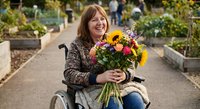 Smiling middle-aged Caucasian woman in wheelchair holding colorful flower bouquet with sunflowers in community garden setting for disability awareness campaigns.