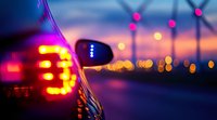 Car side mirror with red brake lights and blurred wind turbines silhouettes against dramatic sunset sky creating atmospheric bokeh effect for environmental concept.