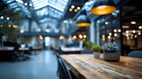 Modern restaurant interior with wooden table in foreground and blurred dining area background featuring pendant lights and glass ceiling architecture.